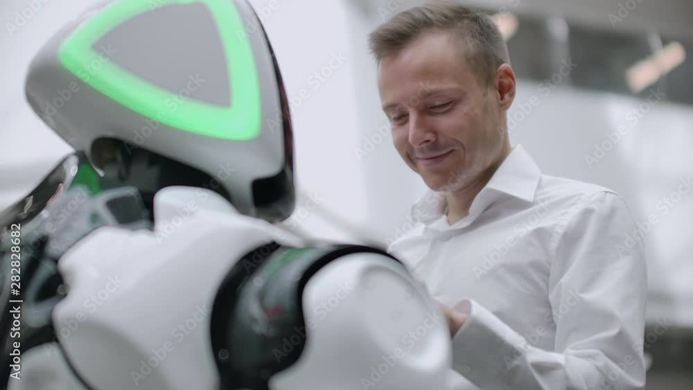 A man stands with a robot bot and asks him questions and asks for help by clicking on the screen on the robot body. A man stands with a robot bot and asks him questions and asks for help by clicking on the screen on the robot body.