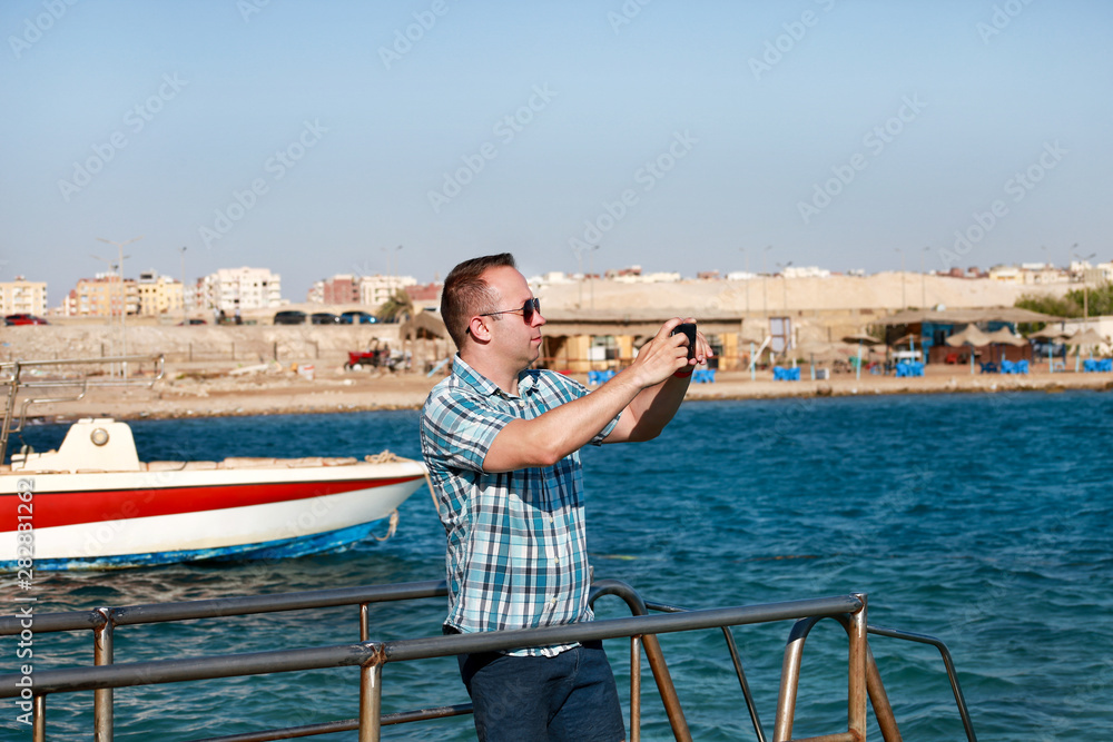 Tourist and traveler on beach pier is taking pictures using camera his smart phone of sea coast, environment and nature landscape. Man with sunglasses on vacation in hotel resort on beach platform.