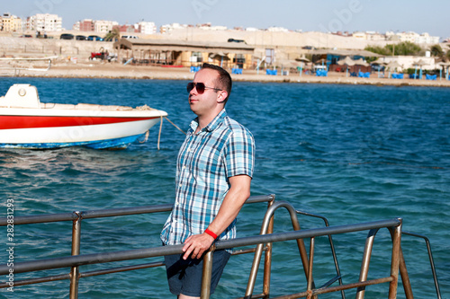 Portrait of tourist and traveler is posing and walking on beach pier, enjoys in sun and view of sea landscape. Man with sunglasses on vacation in summertime in hotel resort on sea jetty platform.