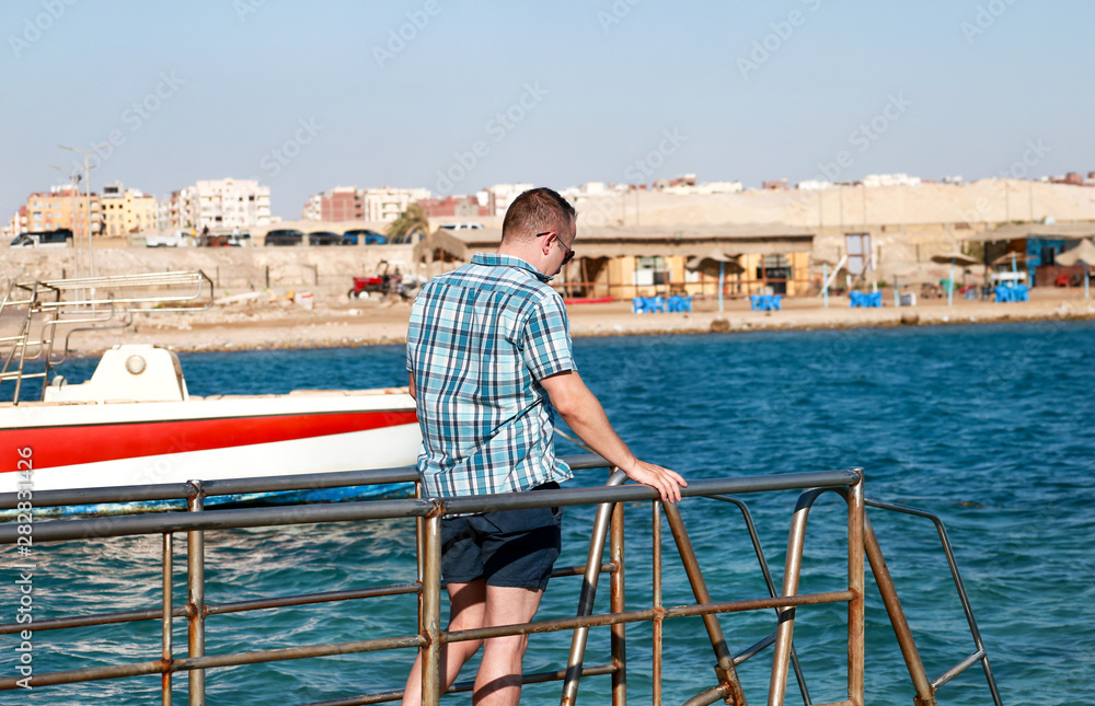 Fototapeta premium Portrait of tourist and traveler is posing and walking on beach pier, enjoys in sun and view of sea landscape. Man with sunglasses on vacation in summertime in hotel resort on sea jetty platform.