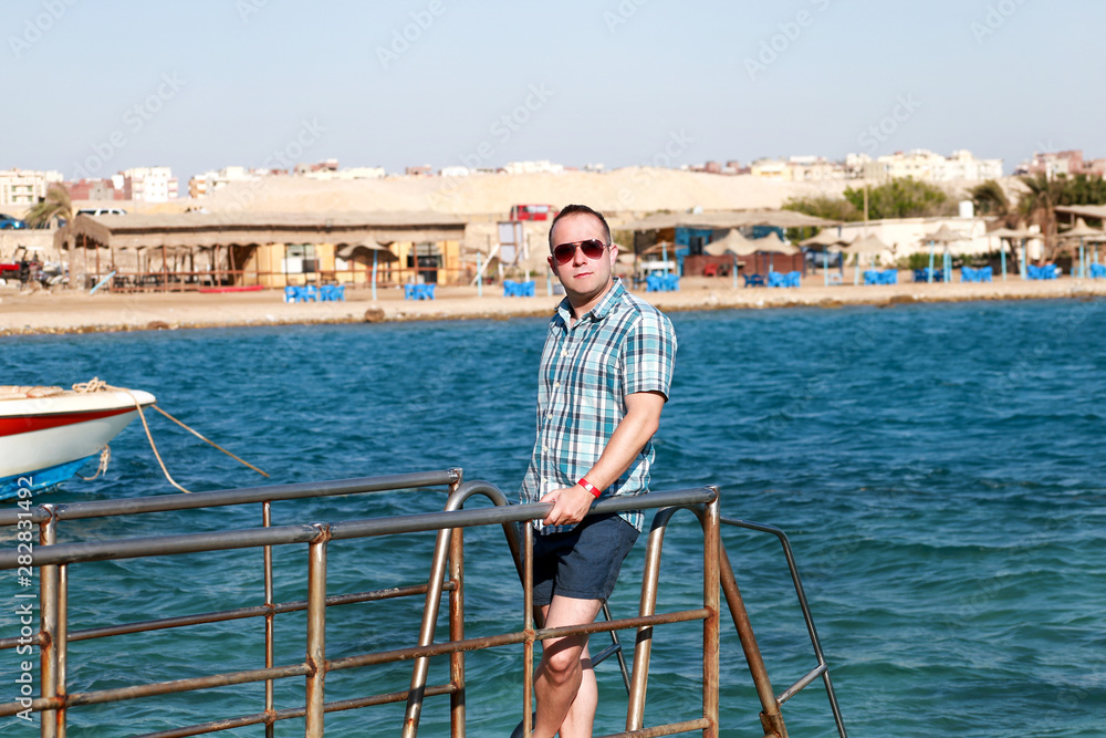 Portrait of tourist and traveler is posing and walking on beach pier, enjoys in sun and view of sea landscape. Man with sunglasses on vacation in summertime in hotel resort on sea jetty platform.