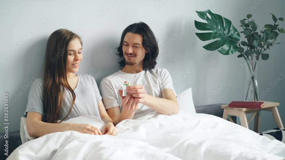 Loving guy is making proposal to girlfriend in bed kissing giving ring in box, woman is saying yes. Relationship, new family and happy young people concept. Loving guy is making proposal to girlfriend in bed kissing giving ring in box, woman is saying yes. Relationship, new family and happy young people concept.