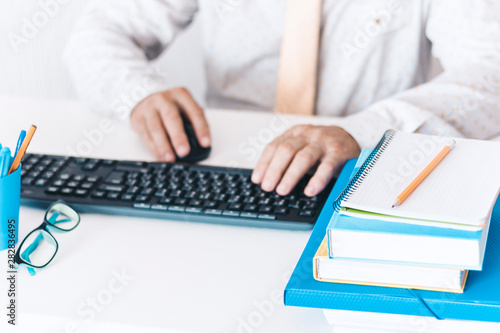 man typing on keyboard laptop computer  plastic blue holder with pencils and pens  stack of books  notebooks  smartphone  glasses on white table  education office concept background.