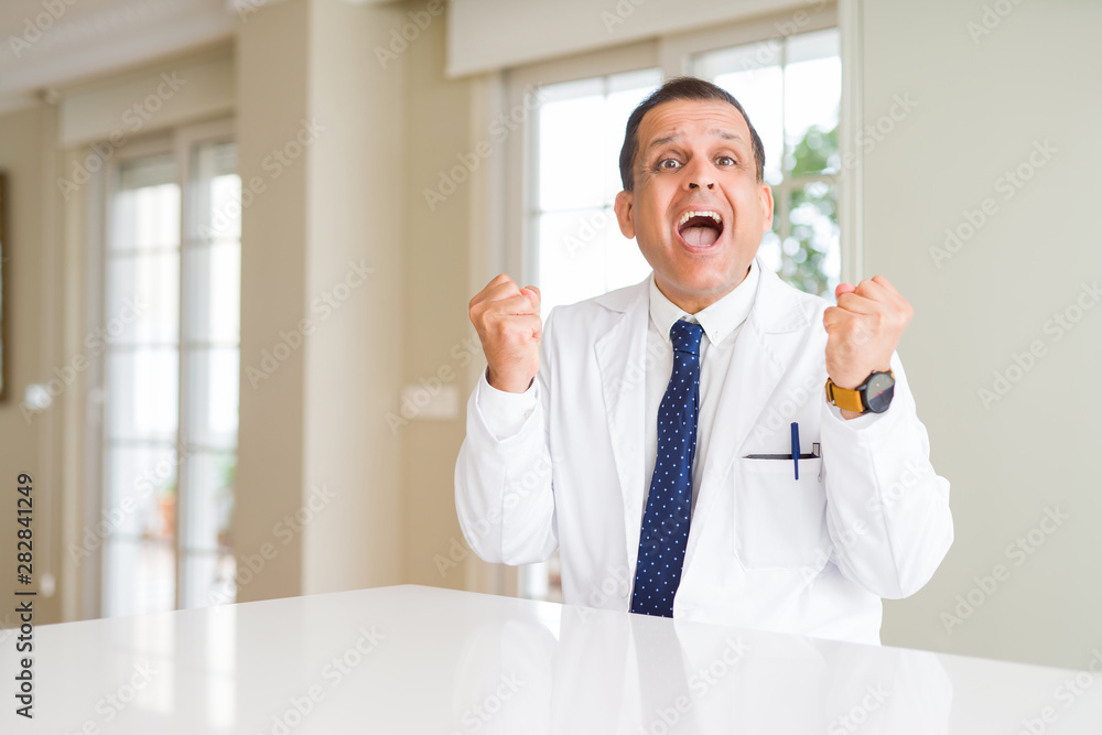 Middle age doctor man wearing medical coat at the clinic celebrating surprised and amazed for success with arms raised and open eyes. Winner concept. Middle age doctor man wearing medical coat at the clinic celebrating surprised and amazed for success with arms raised and open eyes. Winner concept.