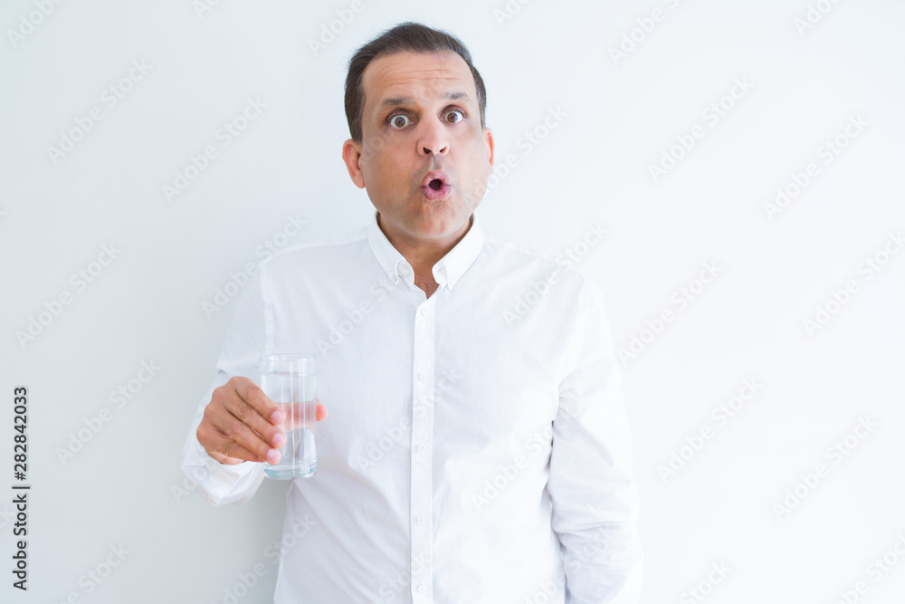 Middle age man drinking a glass of water over white background scared in shock with a surprise face, afraid and excited with fear expression Middle age man drinking a glass of water over white background scared in shock with a surprise face, afraid and excited with fear expression