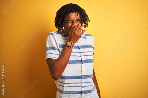 Afro american man with dreadlocks wearing striped shirt over isolated yellow background smelling something stinky and disgusting, intolerable smell, holding breath with fingers on nose. Bad smells © Krakenimages.com