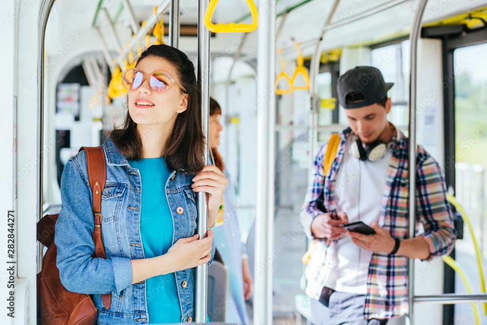 Young adorable joyful woman is standing on the bus using the phone and smiling. - obrazy, fototapety, plakaty 