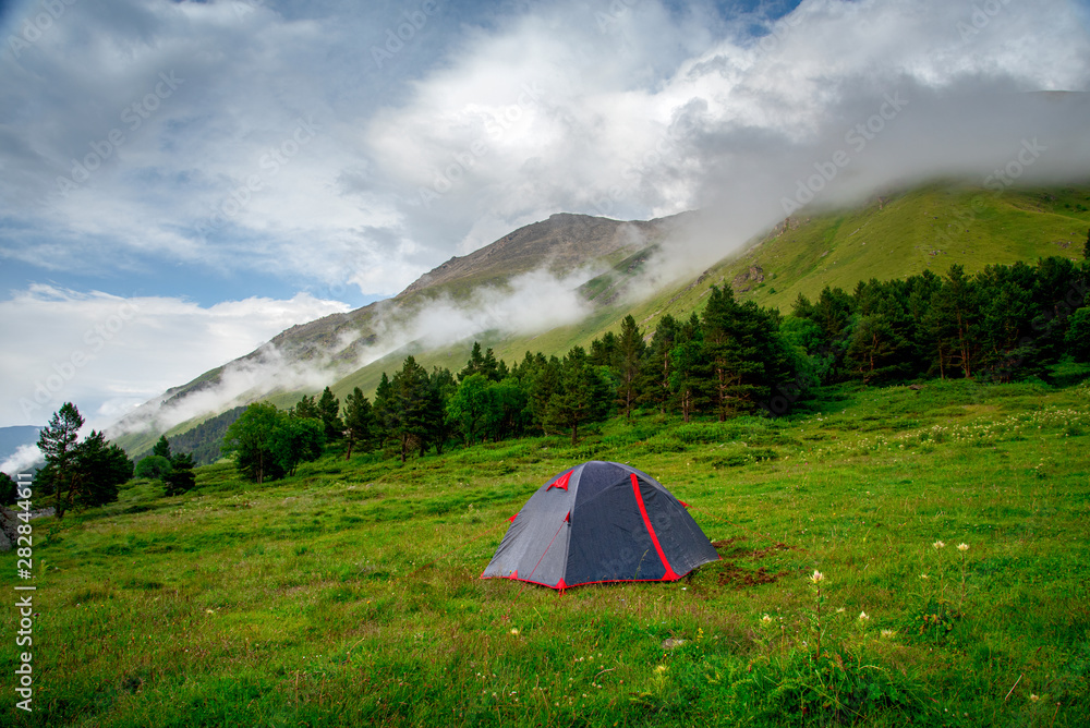 tourist tent stands on a green meadow against the backdrop of mountain slopes and peaks with clouds and blue sky