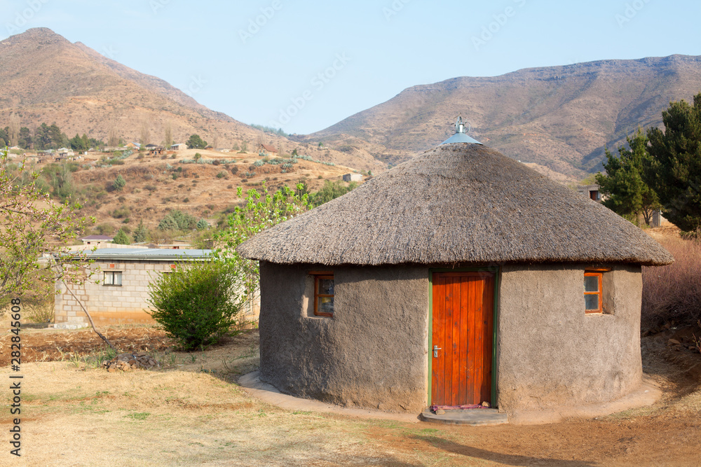 Traditional african round clay house with thatched roof in village, Kingdom of Lesotho, Southern Africa, ethnic basotho people national old home close up, Drakensberg mountains landscape background