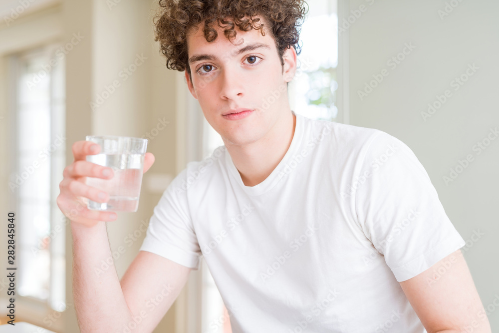 Young man drinking a glass of water at home with a confident expression on smart face thinking serious Young man drinking a glass of water at home with a confident expression on smart face thinking serious