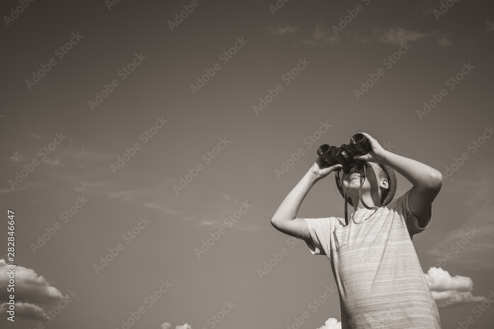 Cute white kid standing alone in summer sunny meadow isolated at clear blue sky background and holding old black binocular in hands. Horizontal black and white sepia color photography. Cute white kid standing alone in summer sunny meadow isolated at clear blue sky background and holding old black binocular in hands. Horizontal black and white sepia color photography.