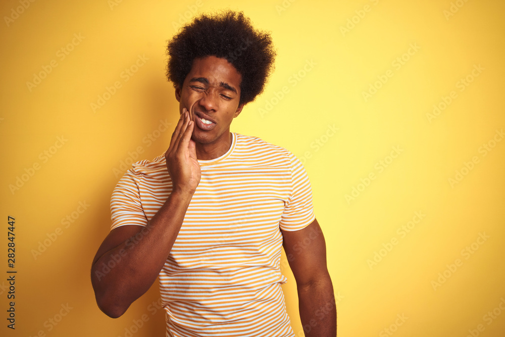 American man with afro hair wearing striped t-shirt standing over isolated yellow background touching mouth with hand with painful expression because of toothache or dental illness on teeth. American man with afro hair wearing striped t-shirt standing over isolated yellow background touching mouth with hand with painful expression because of toothache or dental illness on teeth.