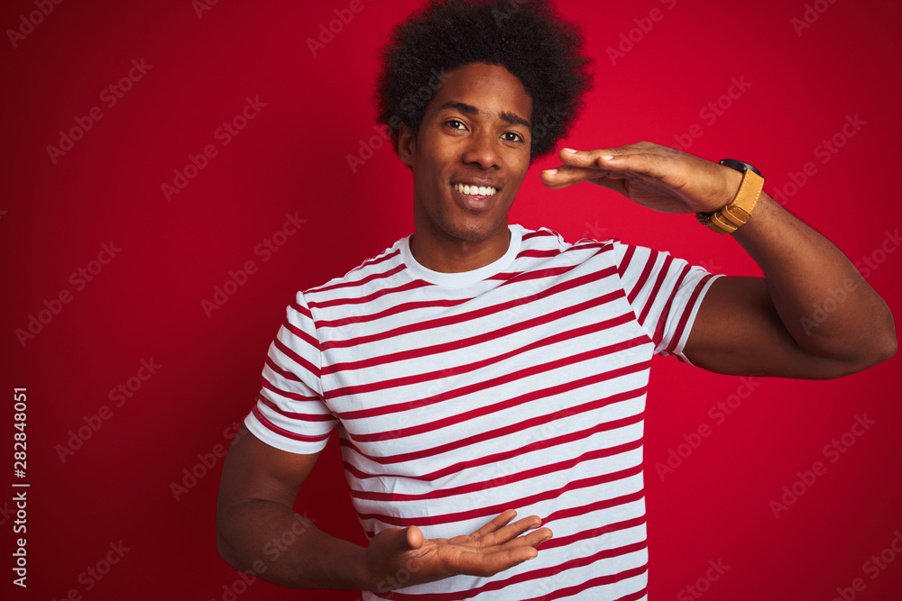 Young african american man with afro hair wearing striped t-shirt over isolated red background gesturing with hands showing big and large size sign, measure symbol. Smiling looking at the camera. Young african american man with afro hair wearing striped t-shirt over isolated red background gesturing with hands showing big and large size sign, measure symbol. Smiling looking at the camera.