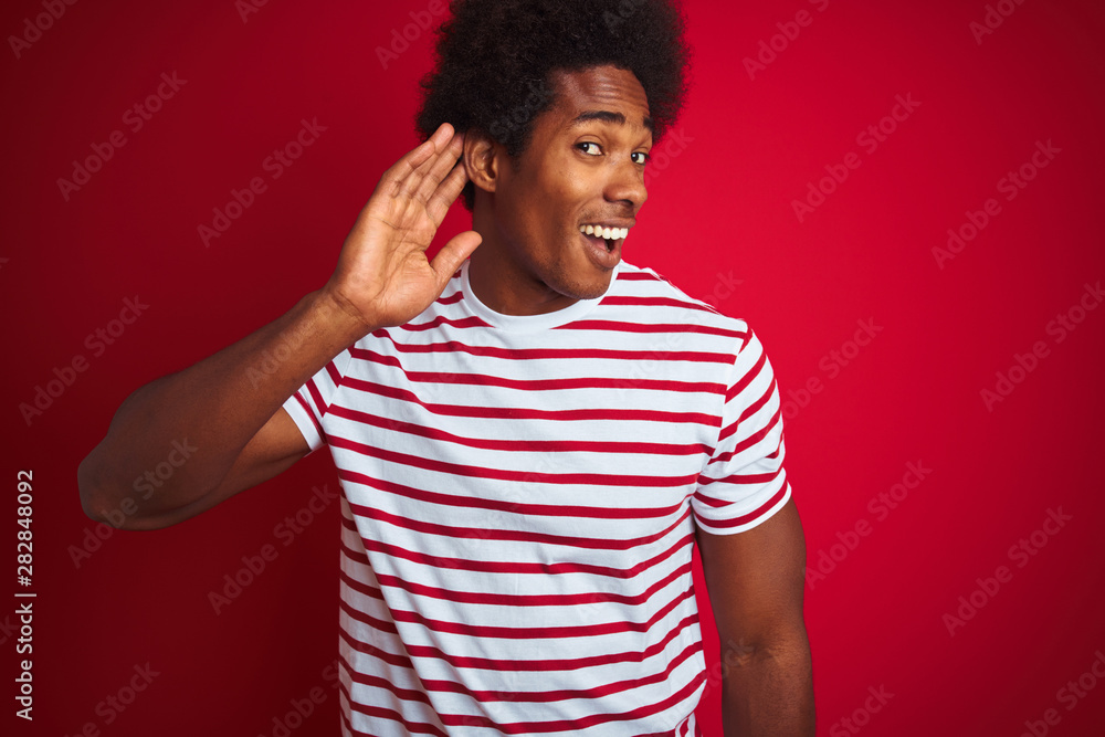 Young african american man with afro hair wearing striped t-shirt over isolated red background smiling with hand over ear listening an hearing to rumor or gossip. Deafness concept. Young african american man with afro hair wearing striped t-shirt over isolated red background smiling with hand over ear listening an hearing to rumor or gossip. Deafness concept.