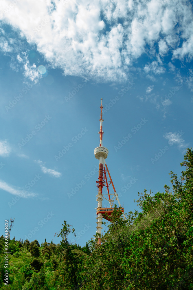 TV tower. Blue sky with clouds. Green trees