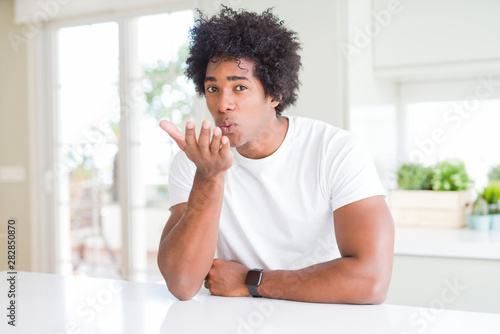 Young african american man wearing casual white t-shirt sitting at home looking at the camera blowing a kiss with hand on air being lovely and sexy. Love expression.