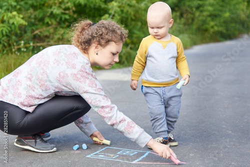 Mother with her little son draws a colorful chalk on asphalt on a summer day. Mom and child activity. © Pavlo