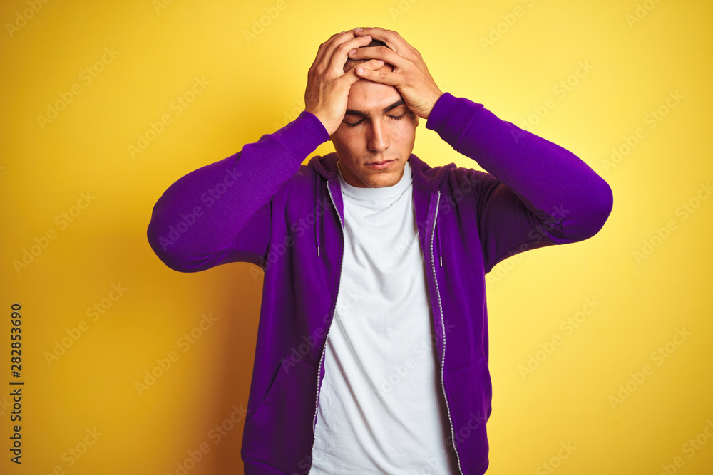 Young handsome man wearing purple sweatshirt over yellow isolated background suffering from headache desperate and stressed because pain and migraine. Hands on head. Young handsome man wearing purple sweatshirt over yellow isolated background suffering from headache desperate and stressed because pain and migraine. Hands on head.