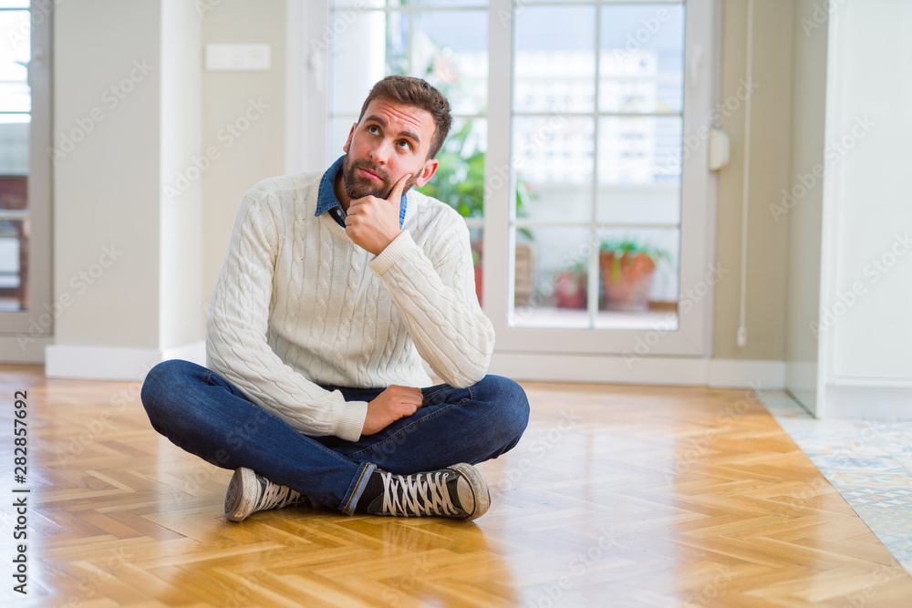 Handsome man wearing casual sweater sitting on the floor at home with hand on chin thinking about question, pensive expression. Smiling with thoughtful face. Doubt concept. Handsome man wearing casual sweater sitting on the floor at home with hand on chin thinking about question, pensive expression. Smiling with thoughtful face. Doubt concept.