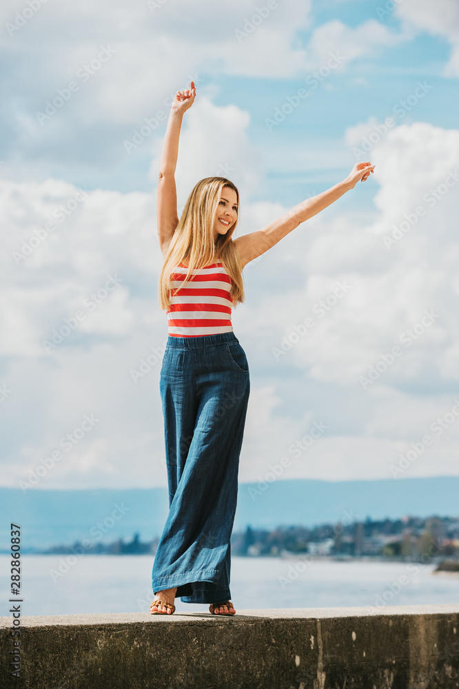 Obraz premium Outdoor fashion portrait of beautiful young woman posing by the lake, wearing red and white stripe vest and denim wide leg jean