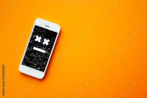 Broken smartphone with cracked destroyed screen on orange background with sad smile. Broken phone service, recovery and repair concept, symbol top view copyspace.