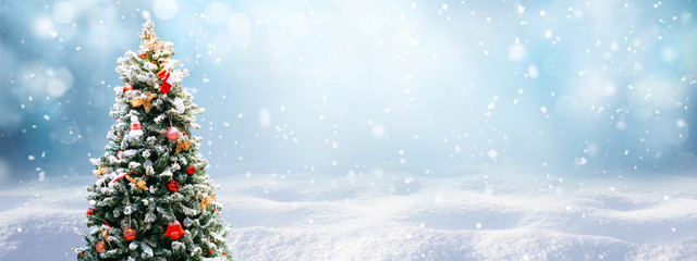 Beautiful Festive Christmas snowy background. Christmas tree decorated with red balls and knitted toys in forest in snowdrifts in snowfall outdoors, banner format, copy space.