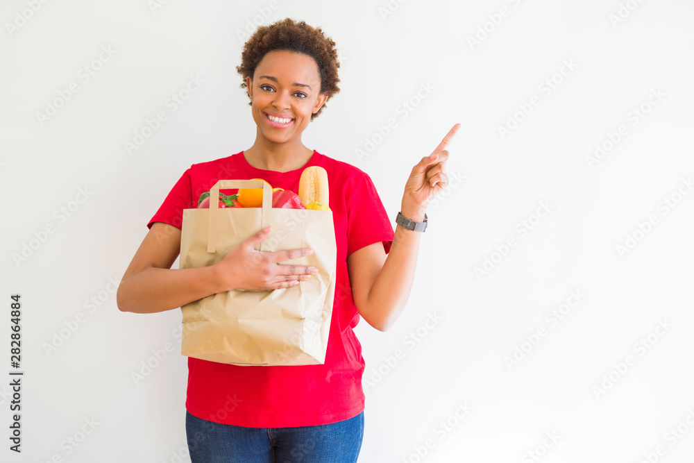 Young african american woman holding paper bag full of fresh groceries very happy pointing with hand and finger to the side Young african american woman holding paper bag full of fresh groceries very happy pointing with hand and finger to the side