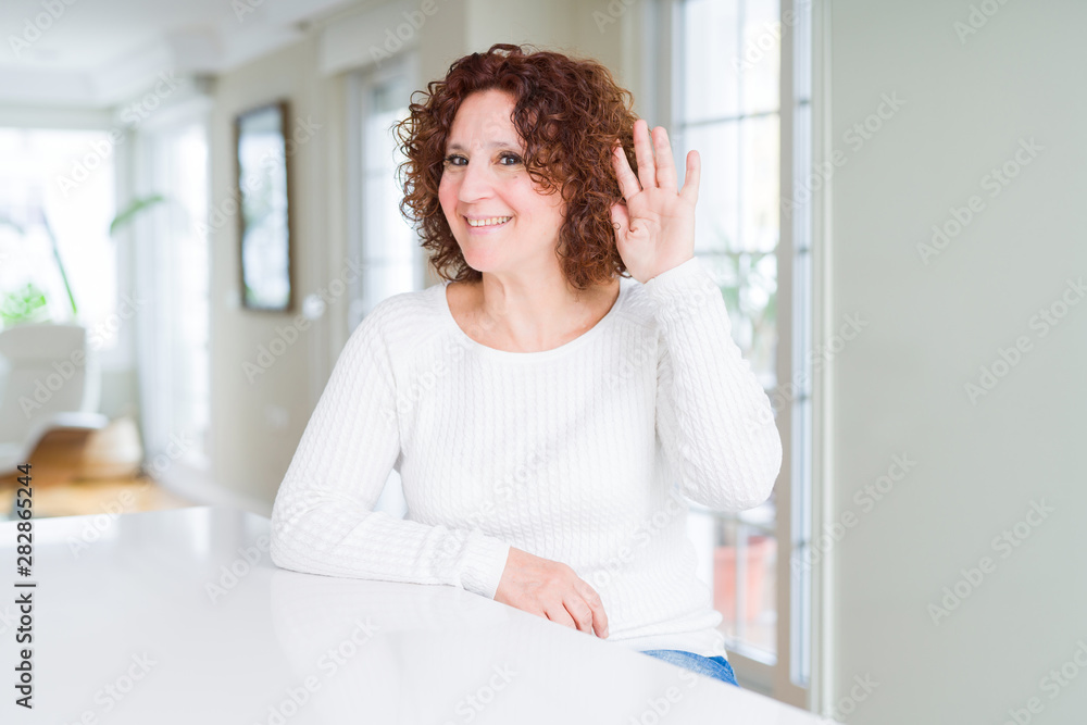 Beautiful senior woman wearing white sweater smiling with hand over ear listening an hearing to rumor or gossip. Deafness concept.