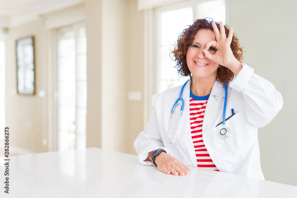 Senior doctor woman wearing medical robe at the clinic doing ok gesture with hand smiling, eye looking through fingers with happy face. Senior doctor woman wearing medical robe at the clinic doing ok gesture with hand smiling, eye looking through fingers with happy face.