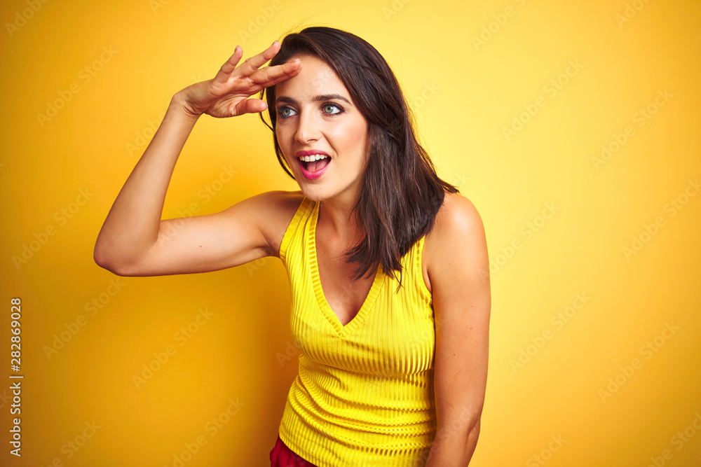 Young beautiful woman wearing t-shirt standing over yellow isolated background very happy and smiling looking far away with hand over head. Searching concept. Young beautiful woman wearing t-shirt standing over yellow isolated background very happy and smiling looking far away with hand over head. Searching concept.