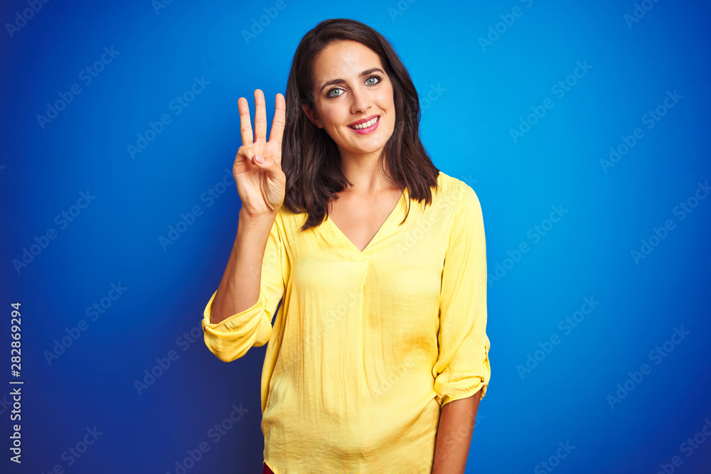 Fototapeta premium Young beautiful woman wearing yellow t-shirt standing over blue isolated background showing and pointing up with fingers number three while smiling confident and happy.