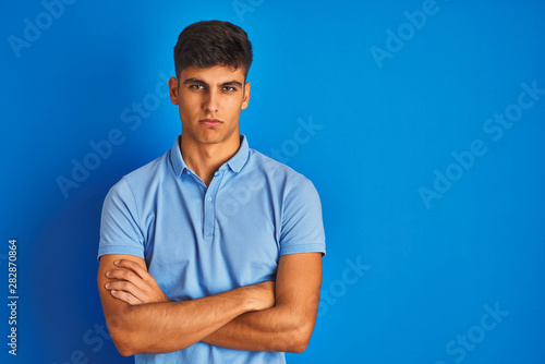 Young indian man wearing casual polo standing over isolated blue background skeptic and nervous, disapproving expression on face with crossed arms. Negative person.