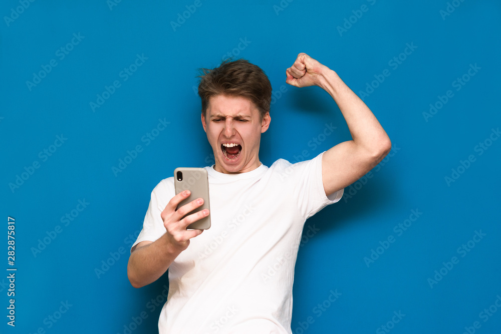 Funny guy in casual clothes is standing on a blue background, takes a selfie on his smartphone and poses for the camera and expresses his emotions. Selfie isolated on blue background Funny guy in casual clothes is standing on a blue background, takes a selfie on his smartphone and poses for the camera and expresses his emotions. Selfie isolated on blue background