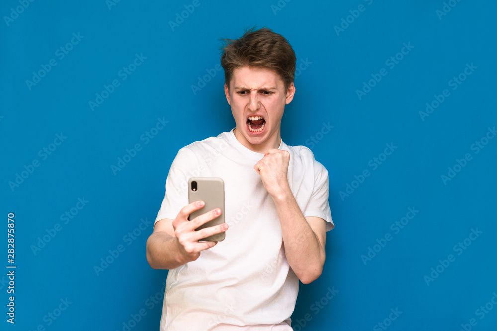 A funny guy in casual clothes is standing on a blue background, takes a selfie on his smartphone and poses for the camera, shows his emotions. Selfie isolated on blue background A funny guy in casual clothes is standing on a blue background, takes a selfie on his smartphone and poses for the camera, shows his emotions. Selfie isolated on blue background