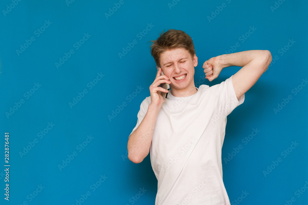 A young man in a white footed coat holds the phone in his ear, emotionally communicates by telephone with his eyes closed with a raised hand holding over his head on a blue background. A young man in a white footed coat holds the phone in his ear, emotionally communicates by telephone with his eyes closed with a raised hand holding over his head on a blue background.