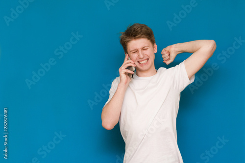 A young man in a white footed coat holds the phone in his ear, emotionally communicates by telephone with his eyes closed with a raised hand holding over his head on a blue background. 
