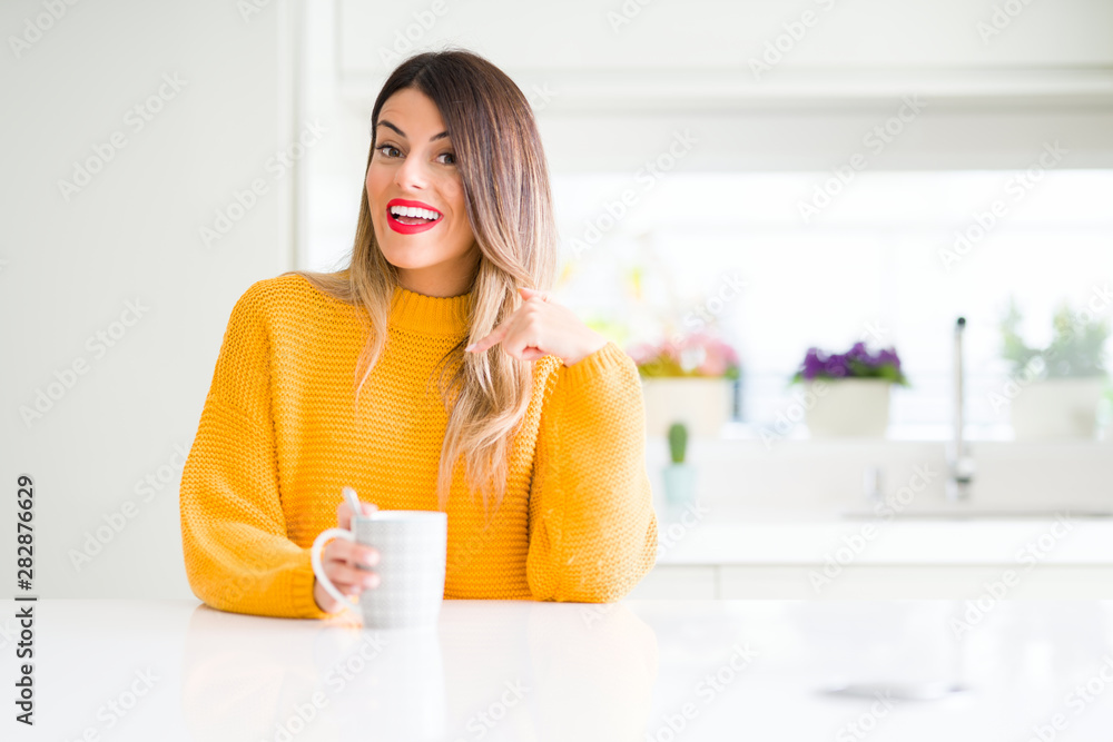 Young beautiful woman drinking a cup of coffee at home with surprise face pointing finger to himself Young beautiful woman drinking a cup of coffee at home with surprise face pointing finger to himself