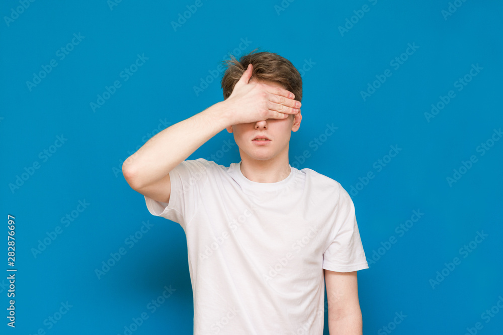 A mysterious man in a white T-shirt covers his face with his hands, against a blue background. Blue background. A mysterious man in a white T-shirt covers his face with his hands, against a blue background. Blue background.