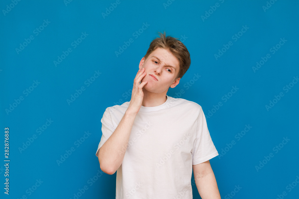 Handsome young man looking down indoors and holding his hand on his chin, standing against a blank backgroundA portrait of an attractive young man shaking his chin and looking down on a blue backgroun Handsome young man looking down indoors and holding his hand on his chin, standing against a blank backgroundA portrait of an attractive young man shaking his chin and looking down on a blue backgroun