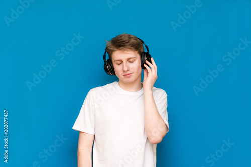 Portrait of an attractive young guy in a white T-shirt stands against the blue wall and listens to music in headphones with closed eyes, enjoys music
