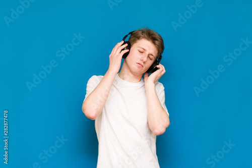 Portrait of an attractive young guy in a white T-shirt is standing against the blue wall and listening to music in headphones with his eyes closed and dancing quietly