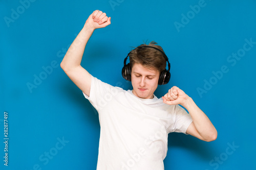 A closeup portrait of a guy who listens to music in headphones on a blue background, looks down and he raised his hands and dances. 
