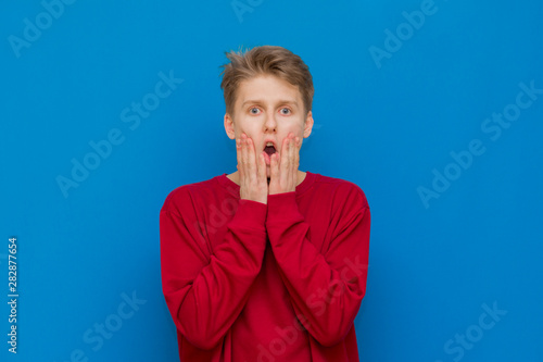 Anxious and insecure young teenage guy with curly hair in shirt biting fingernails looking unsure and nervous as frowning and looking at camera, overthinking as being concerned of things getting worse