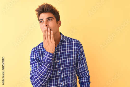 Young handsome man wearing casual shirt standing over isolated yellow background bored yawning tired covering mouth with hand. Restless and sleepiness. © Krakenimages.com