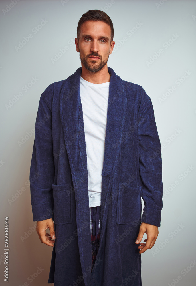 Fototapeta premium Young man wearing comfortable pajamas and robe at morning over isolated background with serious expression on face. Simple and natural looking at the camera.