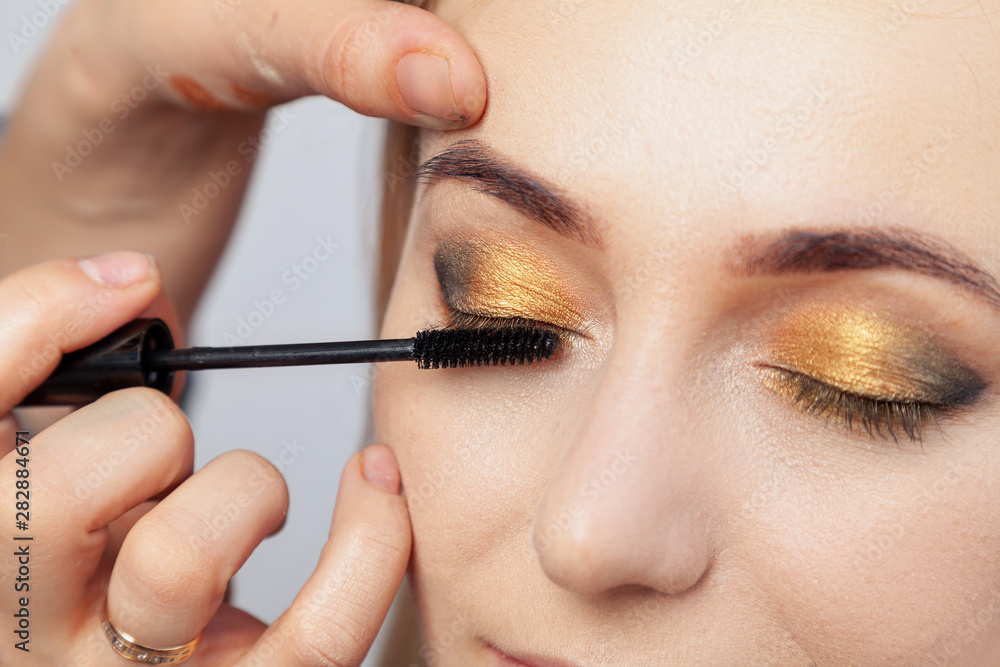 Makeup artist puts on an oriental-style make-up with gold and green shades of a young attractive blonde girl, paints her eyelashes with mascara, hold on the upper eyelid, holding a brush.