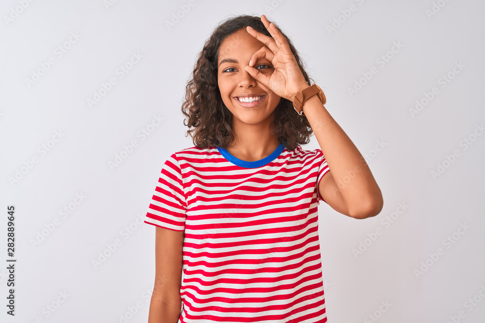Young brazilian woman wearing red striped t-shirt standing over isolated white background doing ok gesture with hand smiling, eye looking through fingers with happy face. Young brazilian woman wearing red striped t-shirt standing over isolated white background doing ok gesture with hand smiling, eye looking through fingers with happy face.