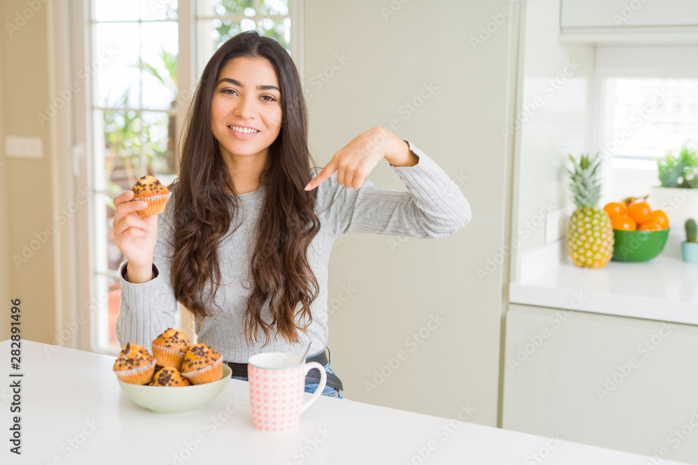 Young woman eating chocolate chips muffins and drinking coffee with surprise face pointing finger to himself Young woman eating chocolate chips muffins and drinking coffee with surprise face pointing finger to himself