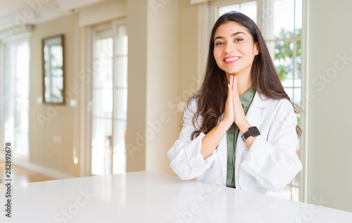 Young woman wearing medical coat at the clinic as therapist or doctor praying with hands together asking for forgiveness smiling confident.