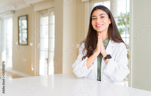 Young woman wearing medical coat at the clinic as therapist or doctor praying with hands together asking for forgiveness smiling confident. Free Download Young woman wearing medical coat at the clinic as therapist or doctor praying with hands together asking for forgiveness smiling confident. - GFXLoad